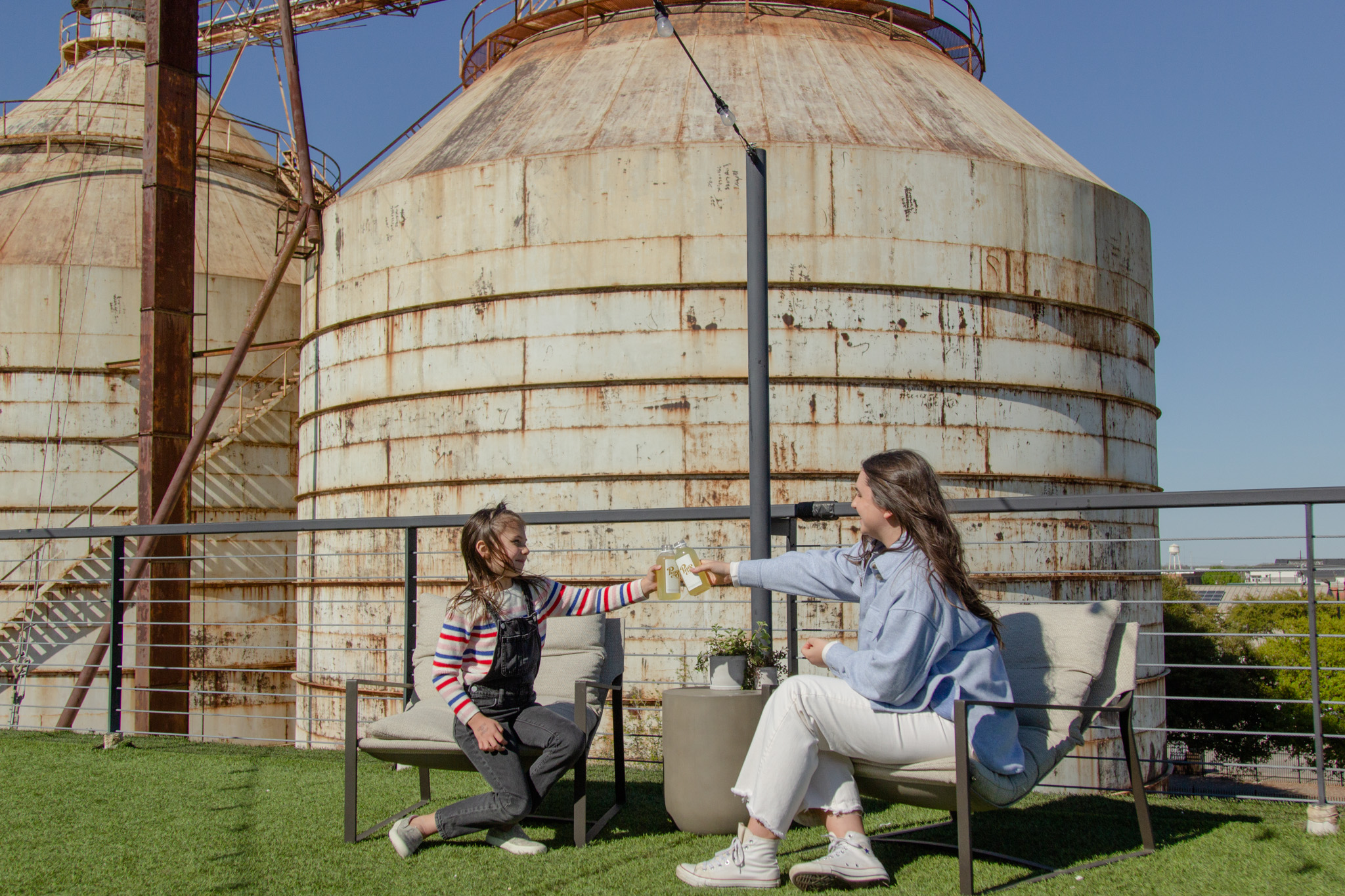 A parent and child seated on the Magnolia Market rooftop lounge, cheers-ing with bottled drinks.