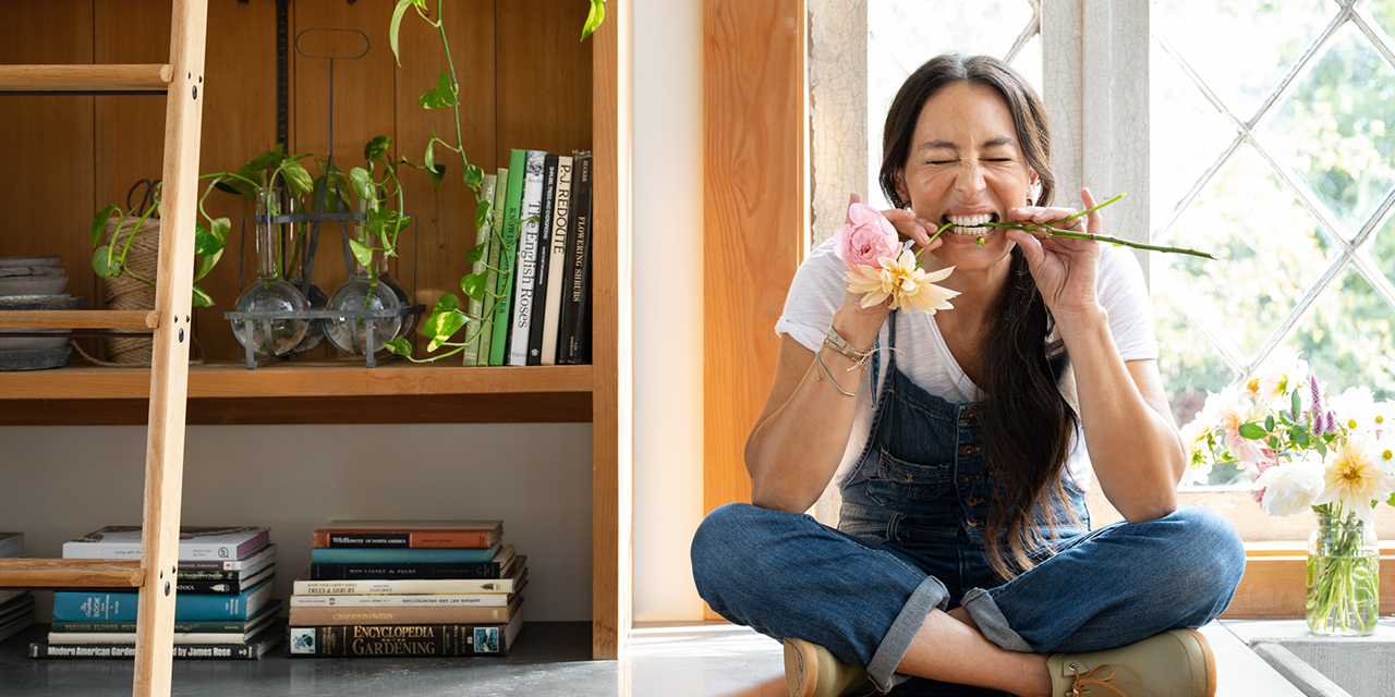 Joanna Gaines sitting on counter in her garden shed with dahilia stems in her mouth
