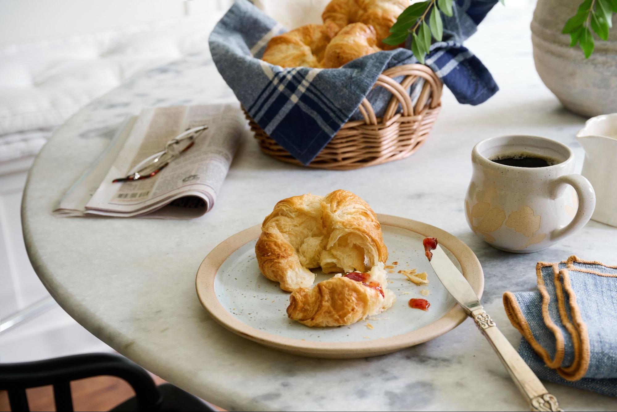 Croissants, newspaper, reading glasses, and a coffee mug sit on a white marble table.