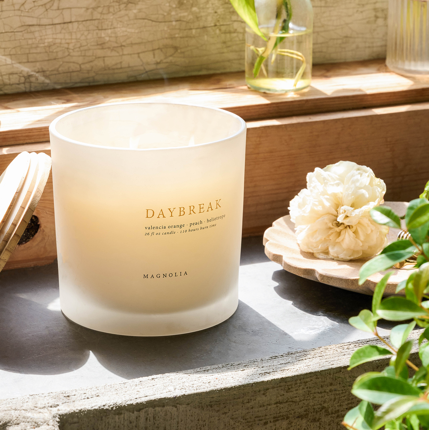 a magnolia daybreak candle sits on a windowsill next to flowers and greenery
