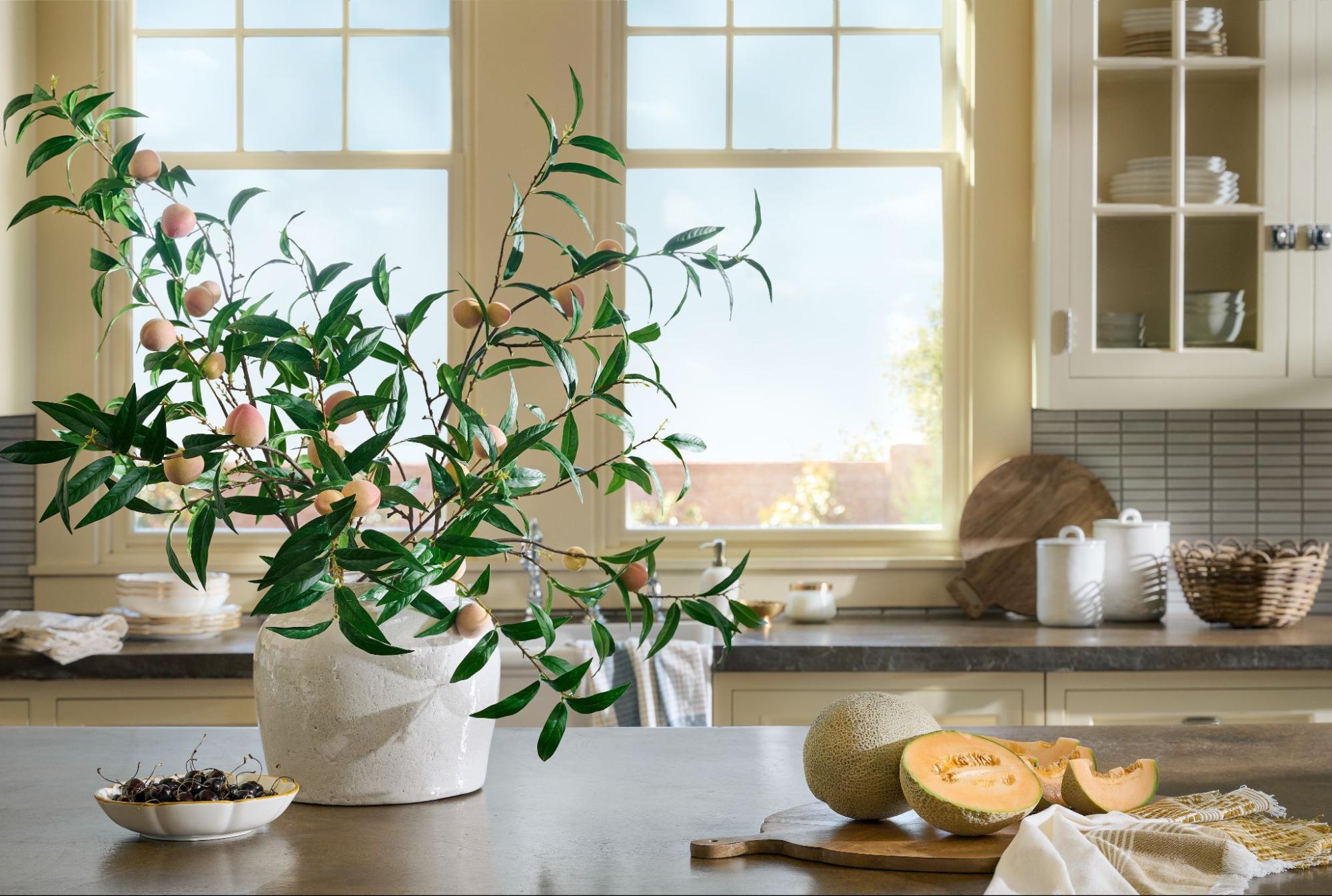 Faux greenery dotted with orange peaches sits in a white vase on a kitchen counter.