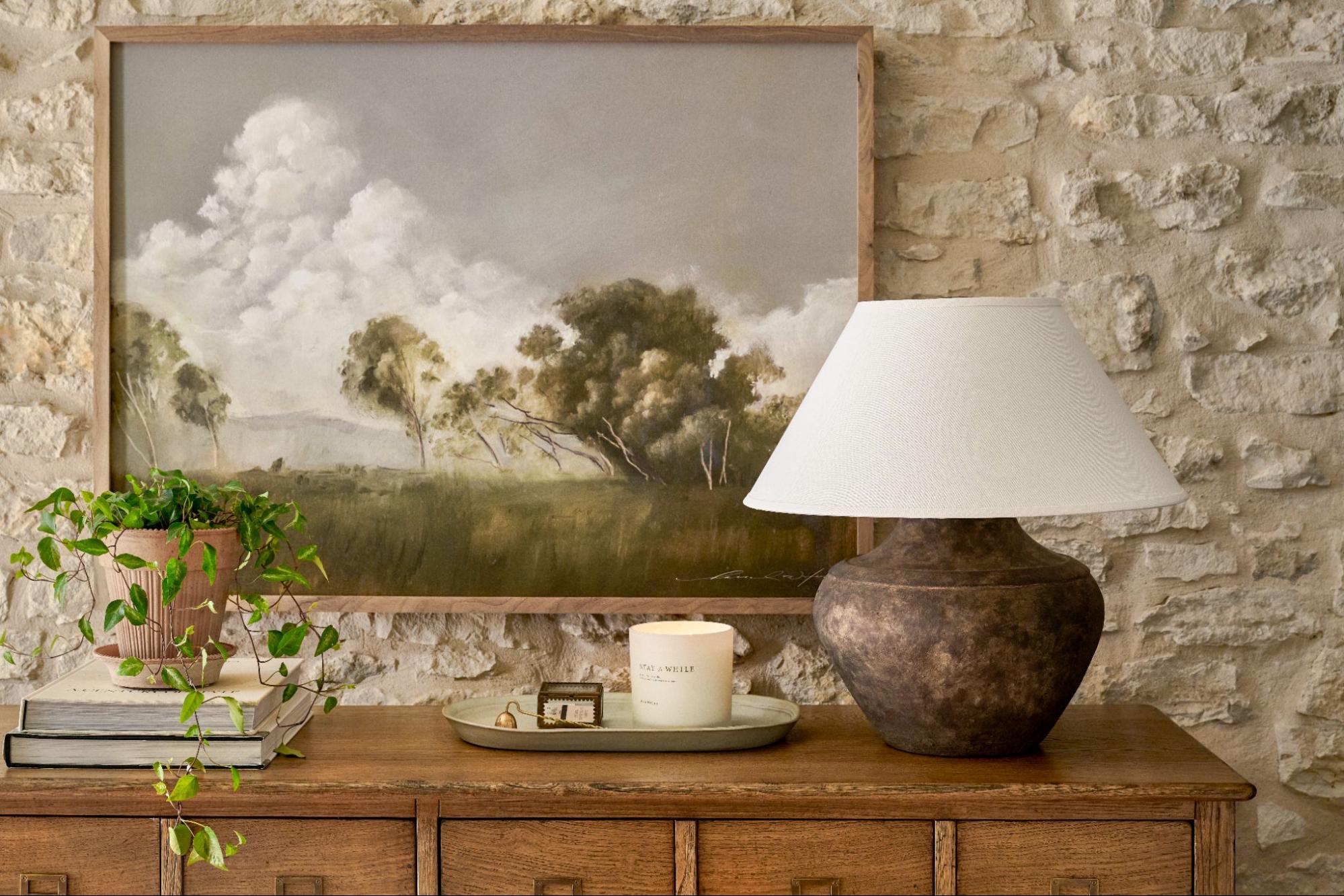 A landscape painting hangs on a stone wall above a styled wooden cabinet. 