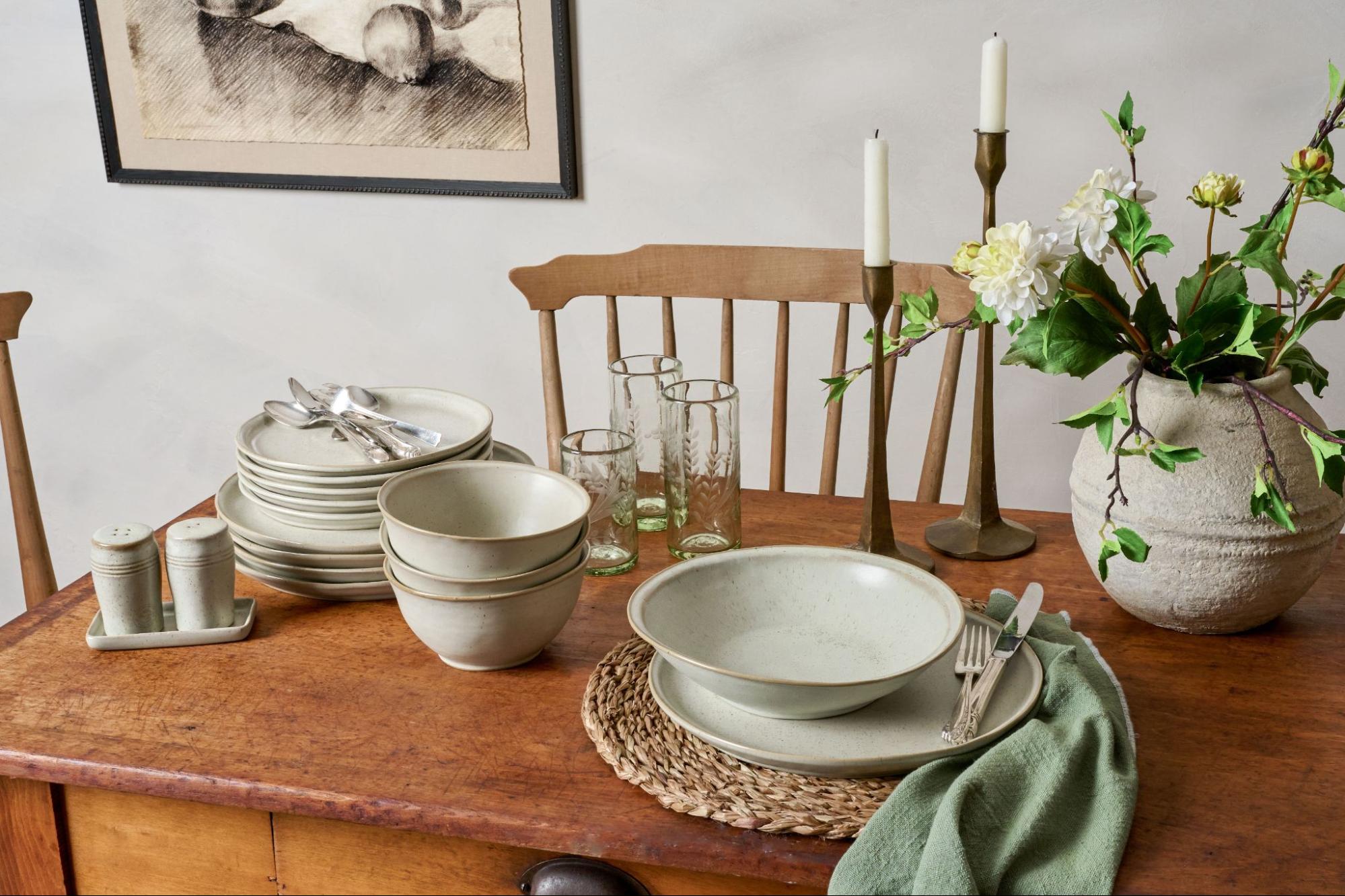 Magnolia tableware sits on a wooden table next to taper candles and a grey vase with faux blooms.