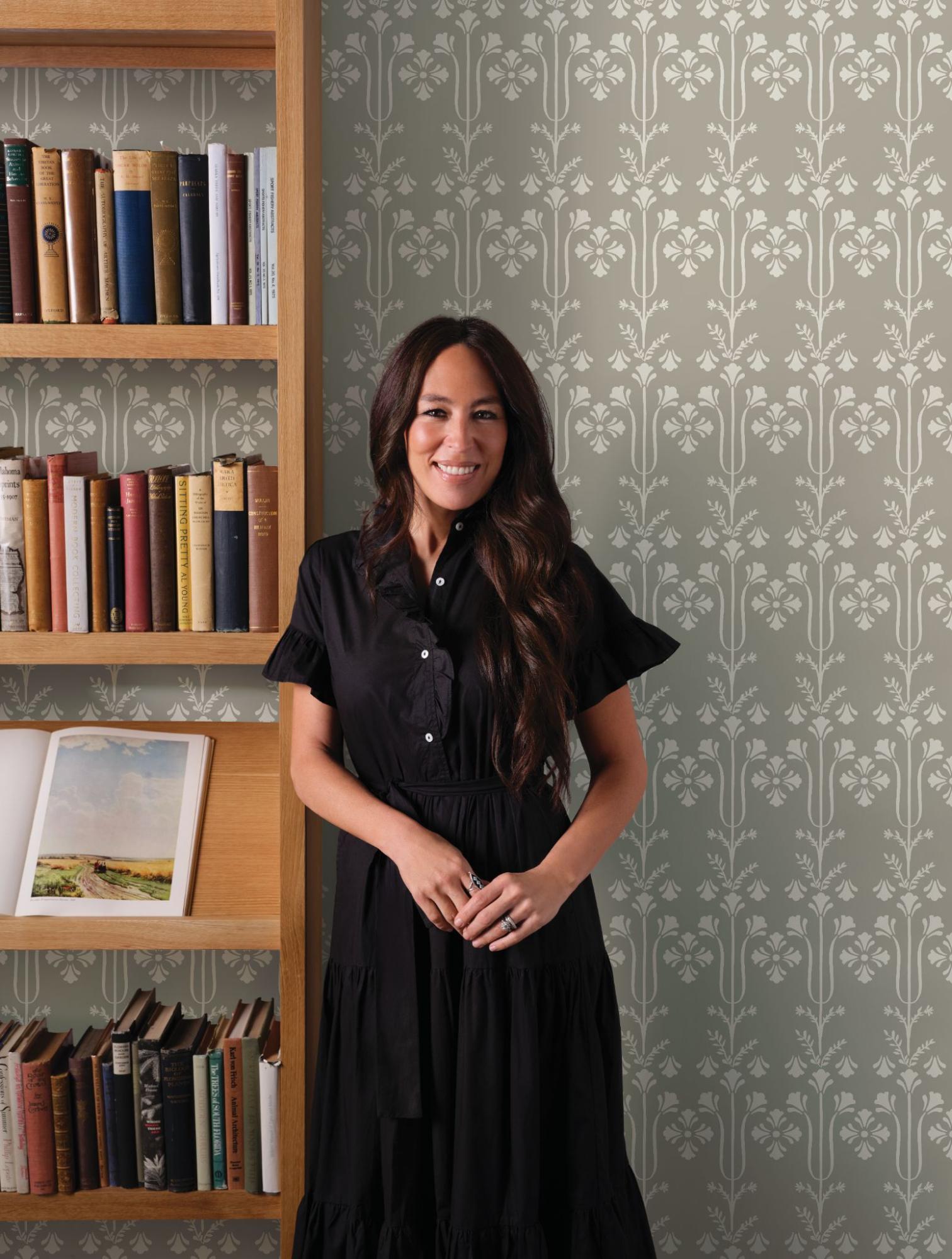 Joanna Gaines smiles in front of a wall with decorative wallpaper and a bookshelf.